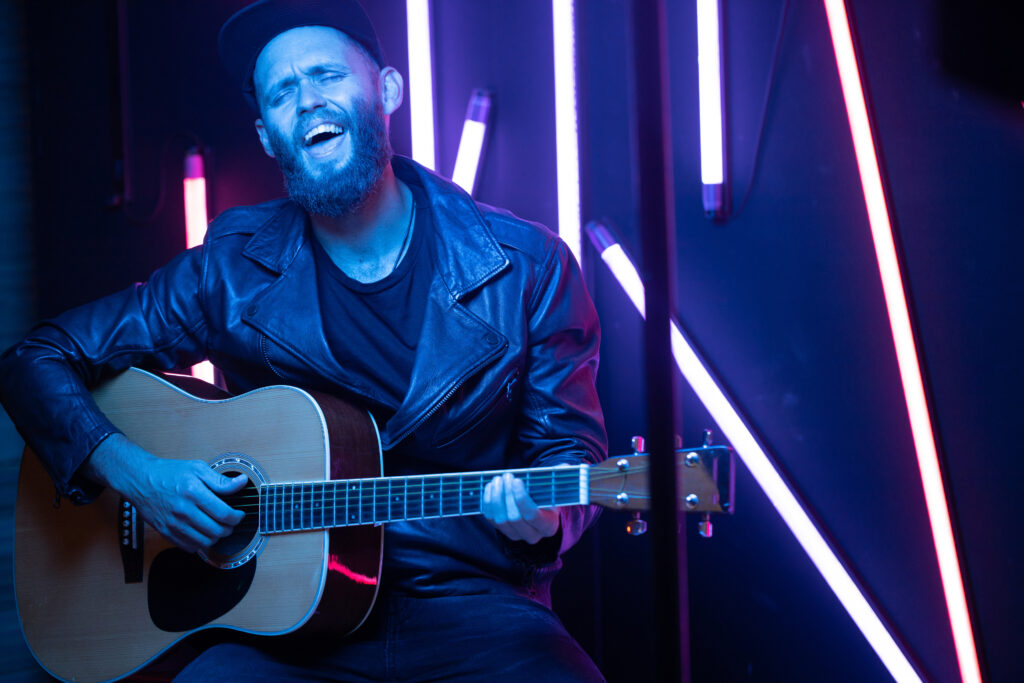 Man singing and playing guitar on a stage with neon lights.